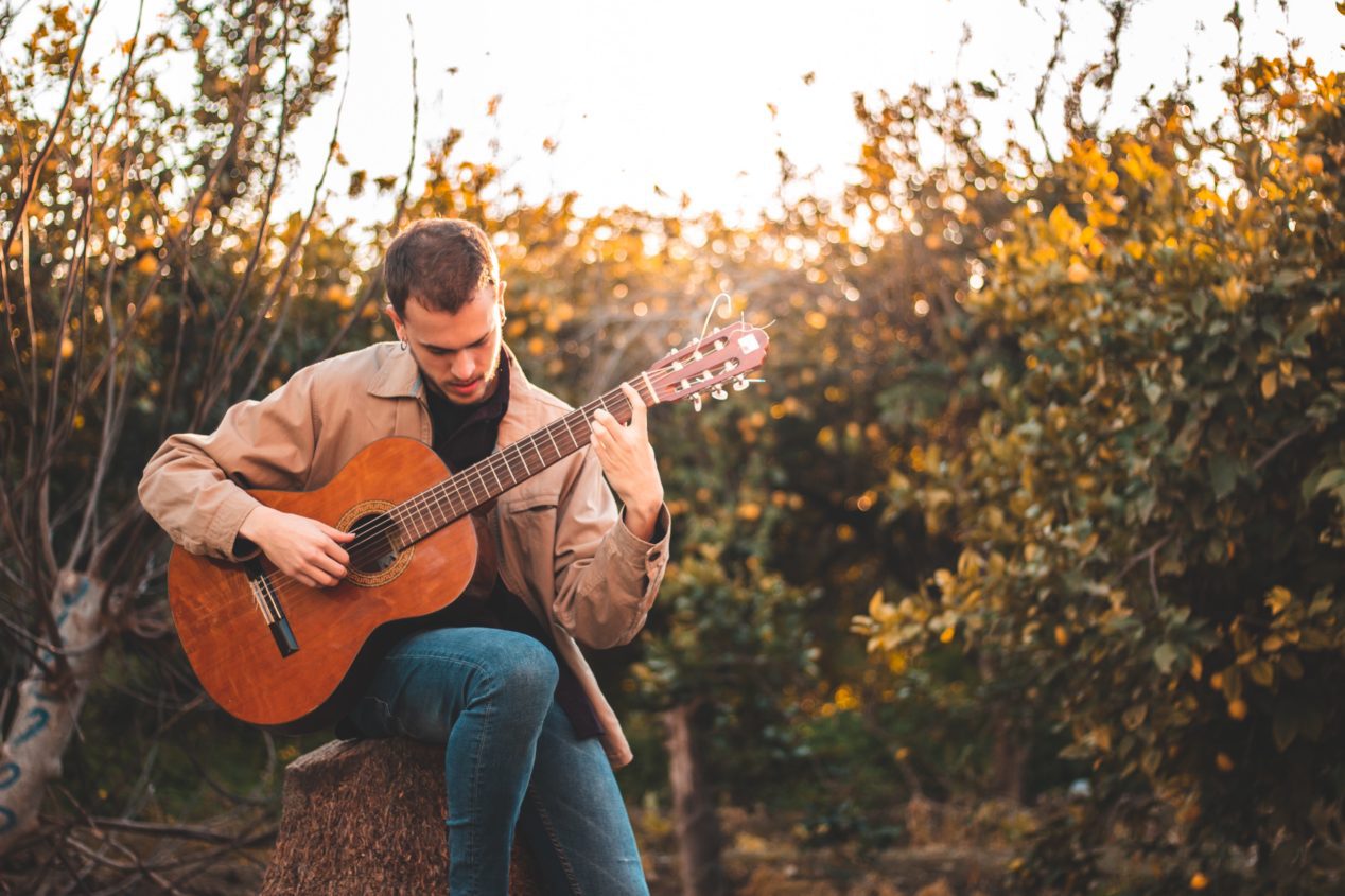 Photo of a man playing a classical guitar outside while sitting on a stump.